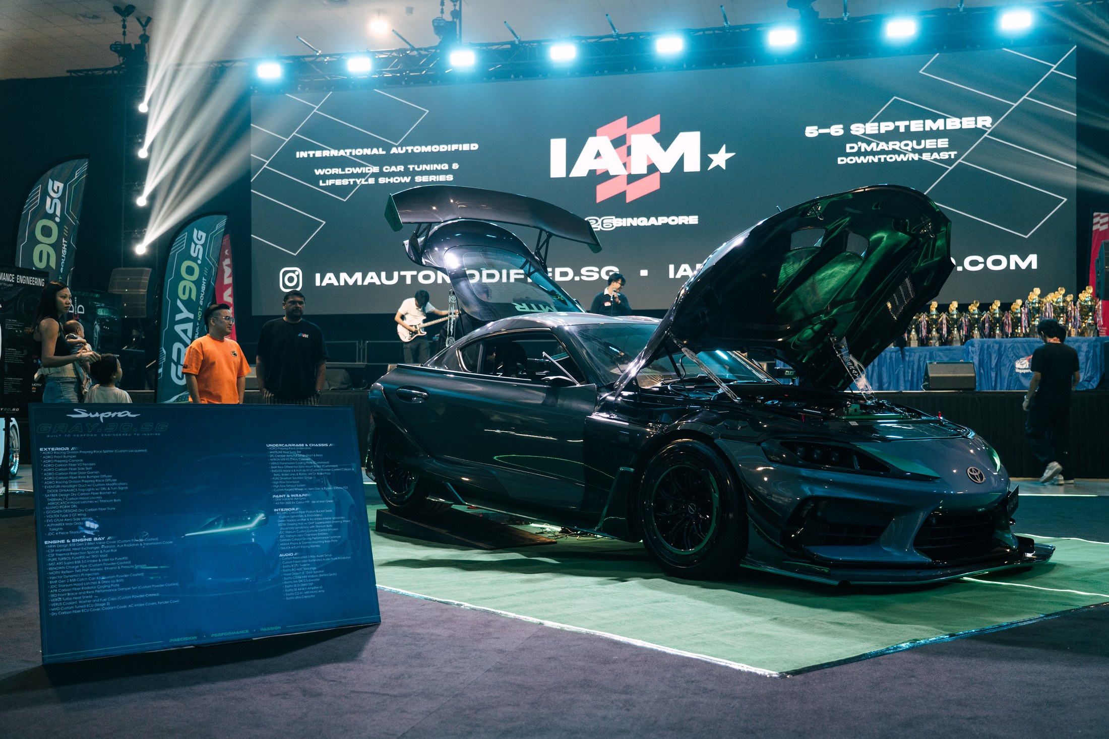 Full reveal at iAM Singapore 2026 — hood and doors up, official show board and trophies in frame, D'Marquee Main Stage behind